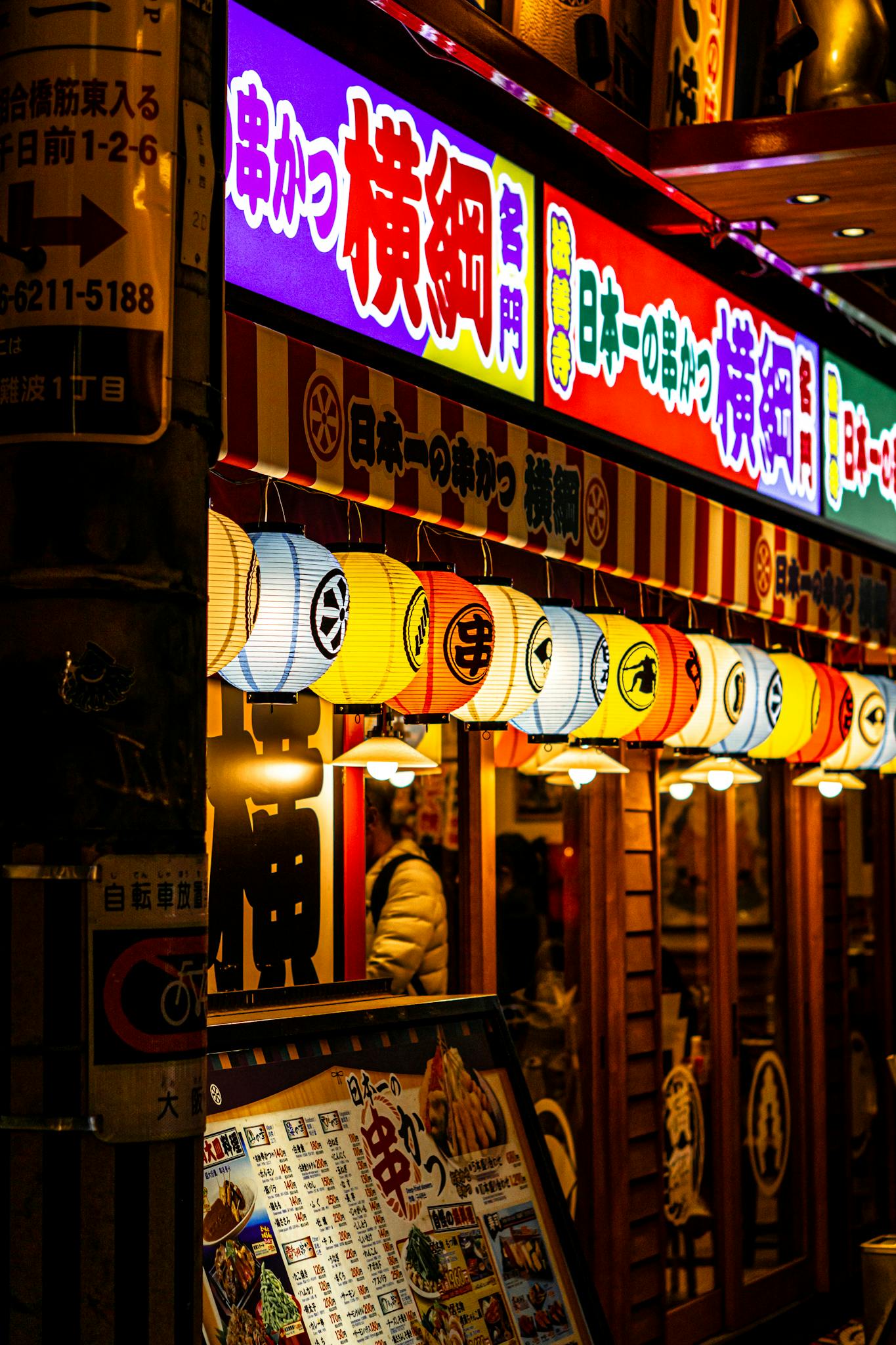 Japanese street food restaurant with vibrant lights and lanterns in a bustling area at night.